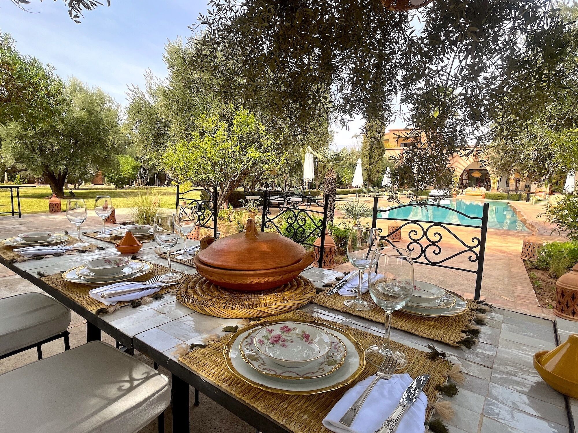 An outdoor dining area with a table set for a meal, surrounded by greenery and a pool in the background.