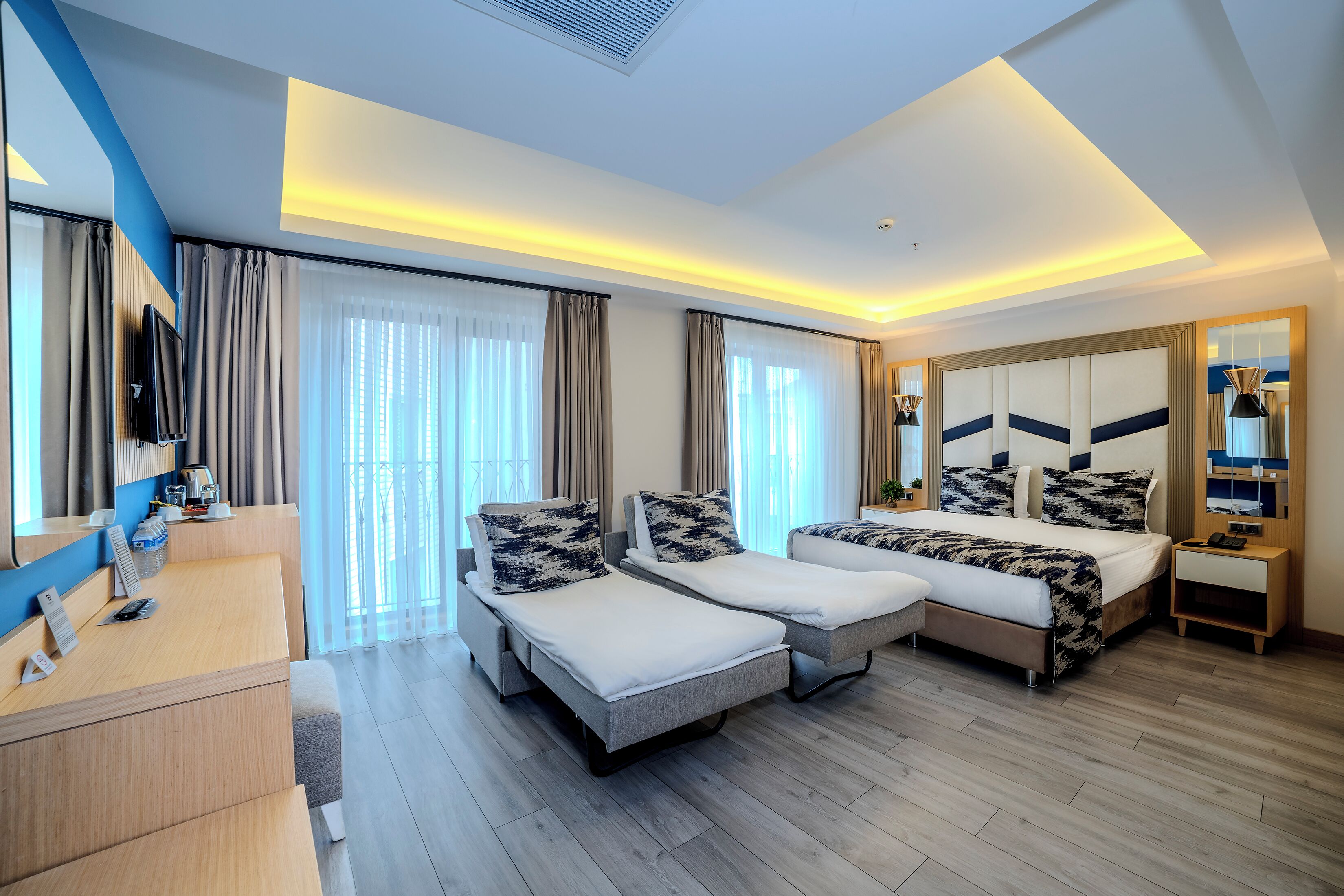A modern hotel room with a bed, two single beds, a desk, and a TV.