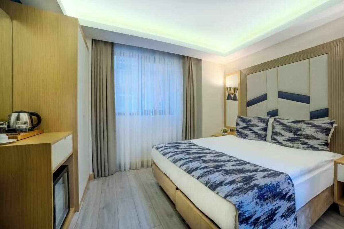 A hotel room with a bed, bedside table, lamp, curtains, and a view of the outdoors.