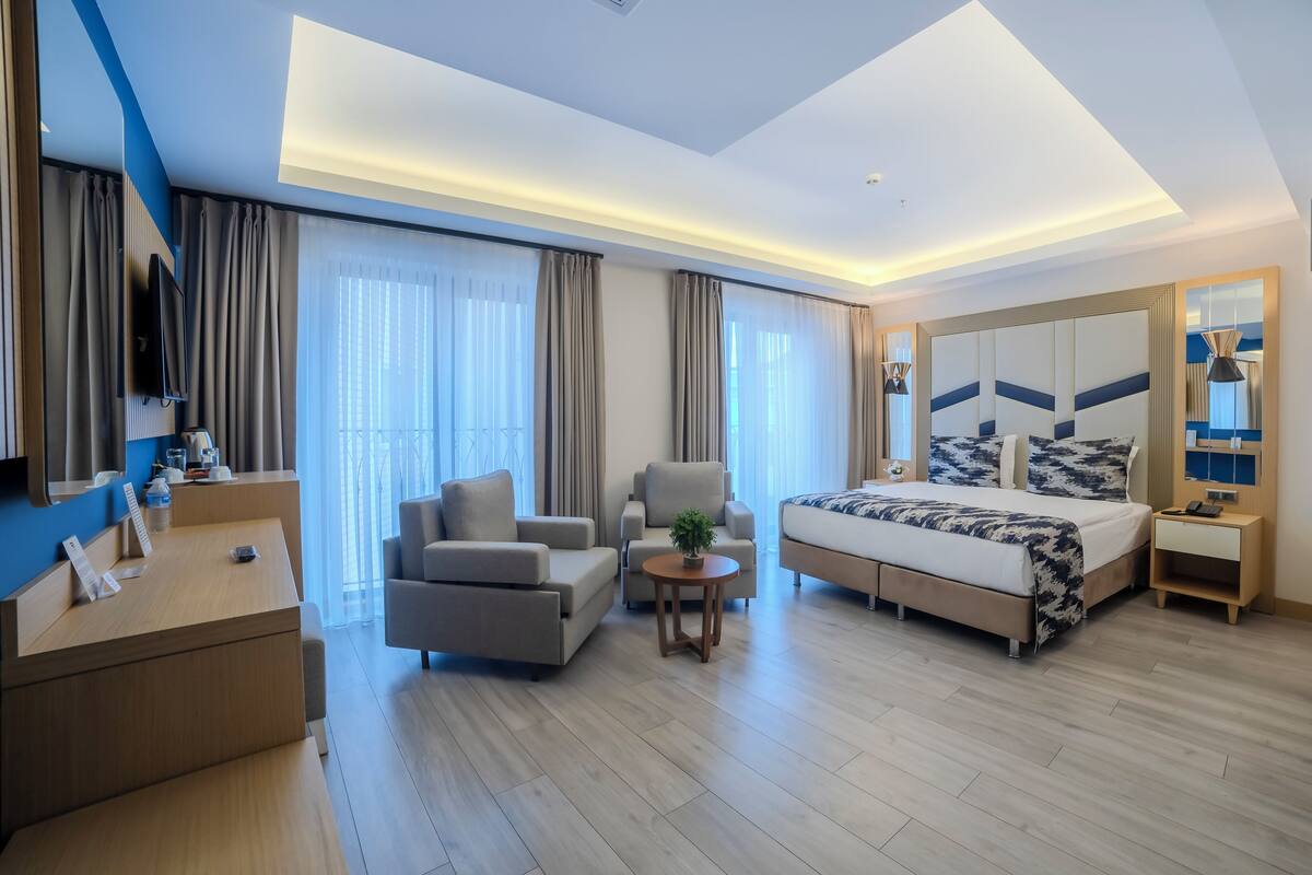 A modern hotel room with a bed, two armchairs, a desk, and a TV.