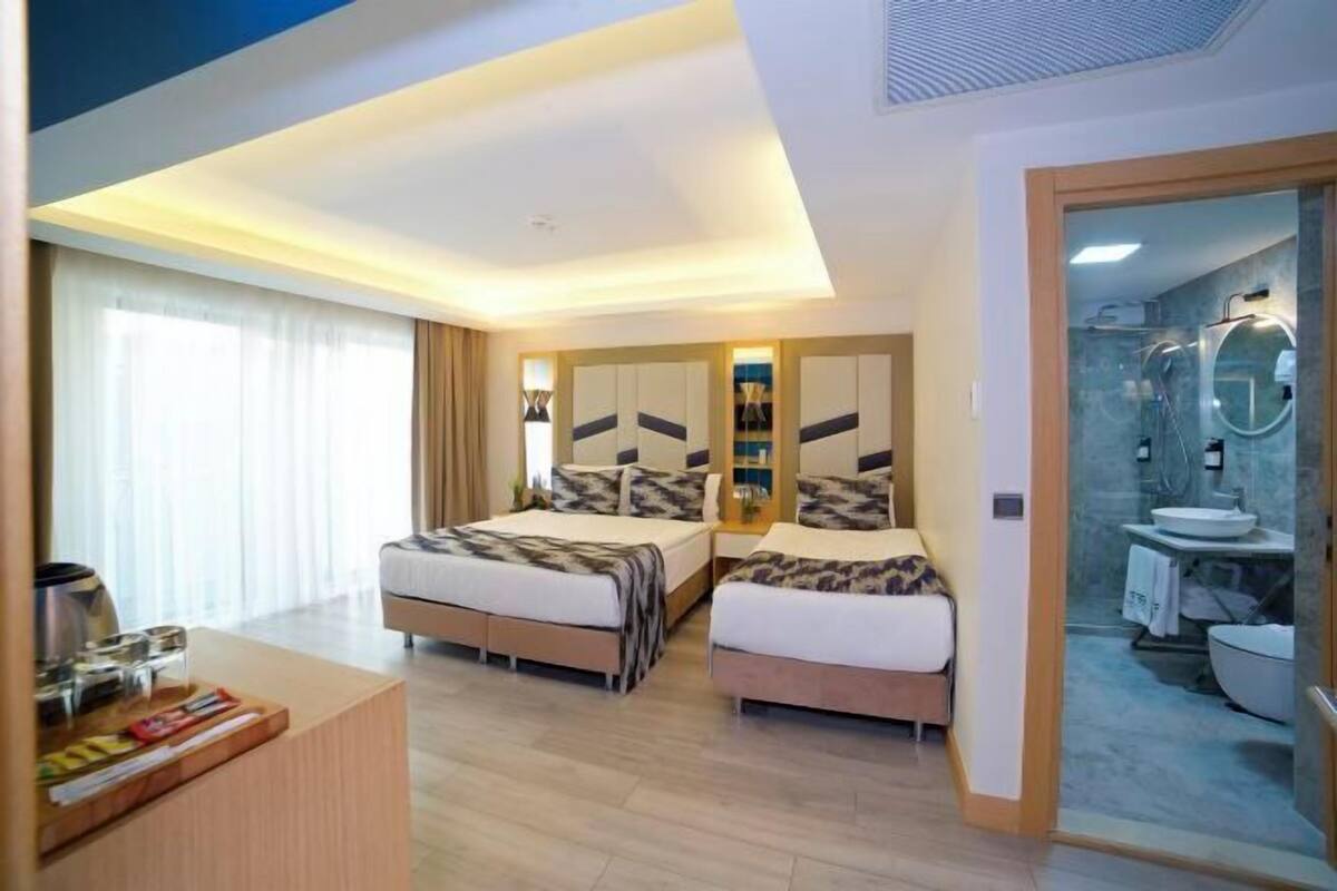 A hotel room with two beds, a desk, a bathroom with a glass shower, and a view of the city.
