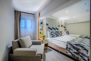 Ein modernes Hotelzimmer mit einer Couch, einem Bett mit gemusterten Bettbezügen, einem Nachttisch mit Pflanze und einem Spiegel mit geometrischer Wandkunst.
