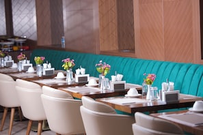 Ein Restaurant mit petrolfarbenen Polstersitzbänken, weißen Stühlen und Tischen, gedeckt mit weißem Geschirr und Blumenvasen.