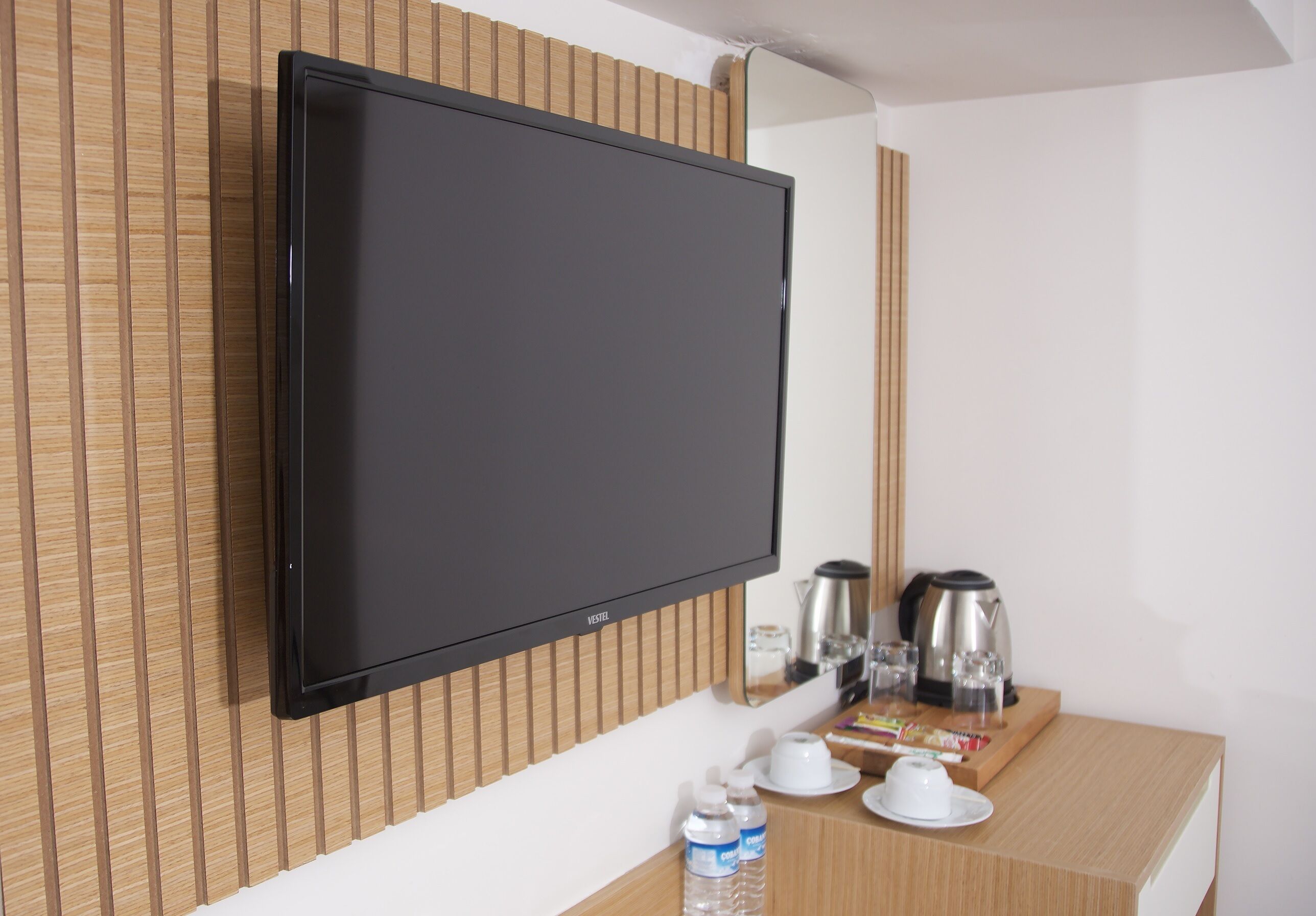 A flat-screen TV mounted on a wooden wall, a coffee maker, a kettle, a tray with cups and a bottle of water on a wooden table.