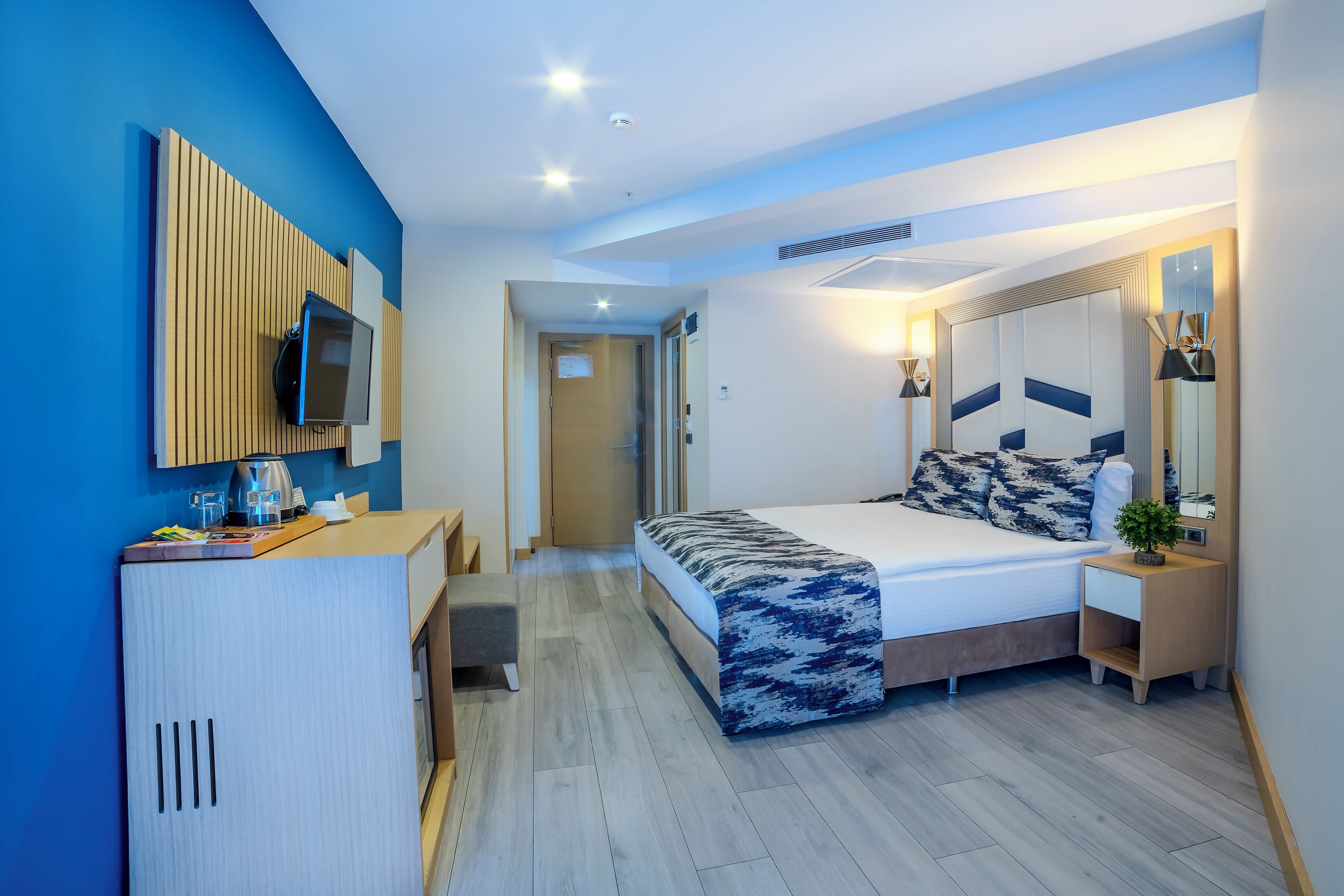 A modern hotel room with a bed, bedside table, TV, and a desk with a chair.