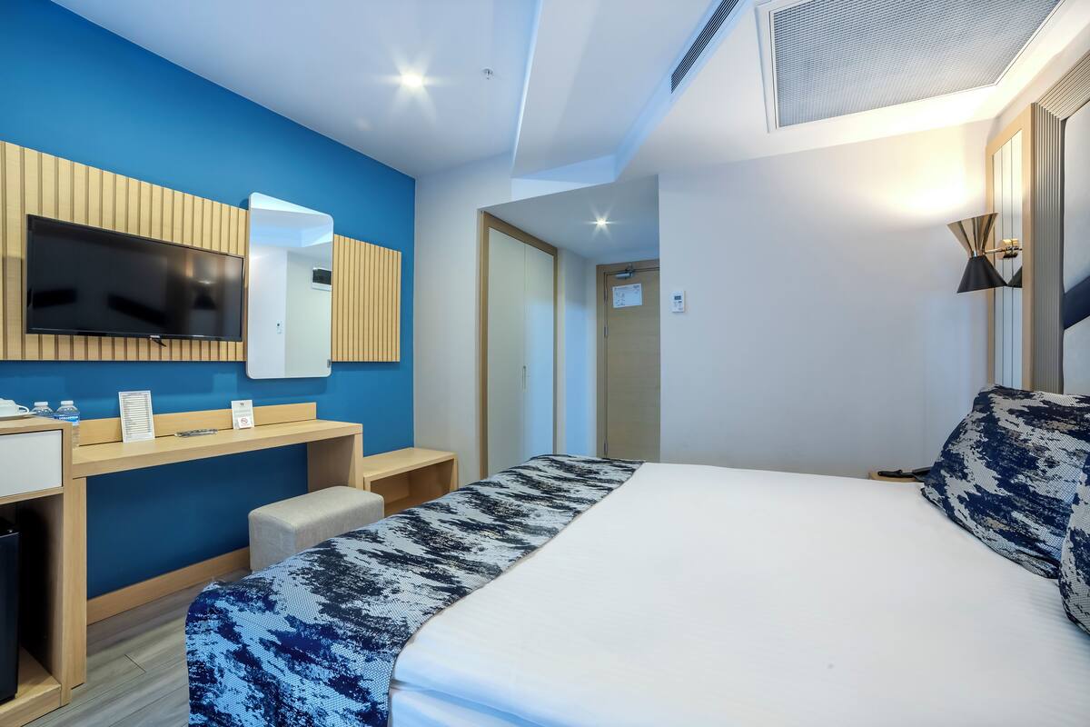 A modern hotel room with a large bed, a desk with a flat-screen TV, a wooden nightstand, and a blue accent wall.