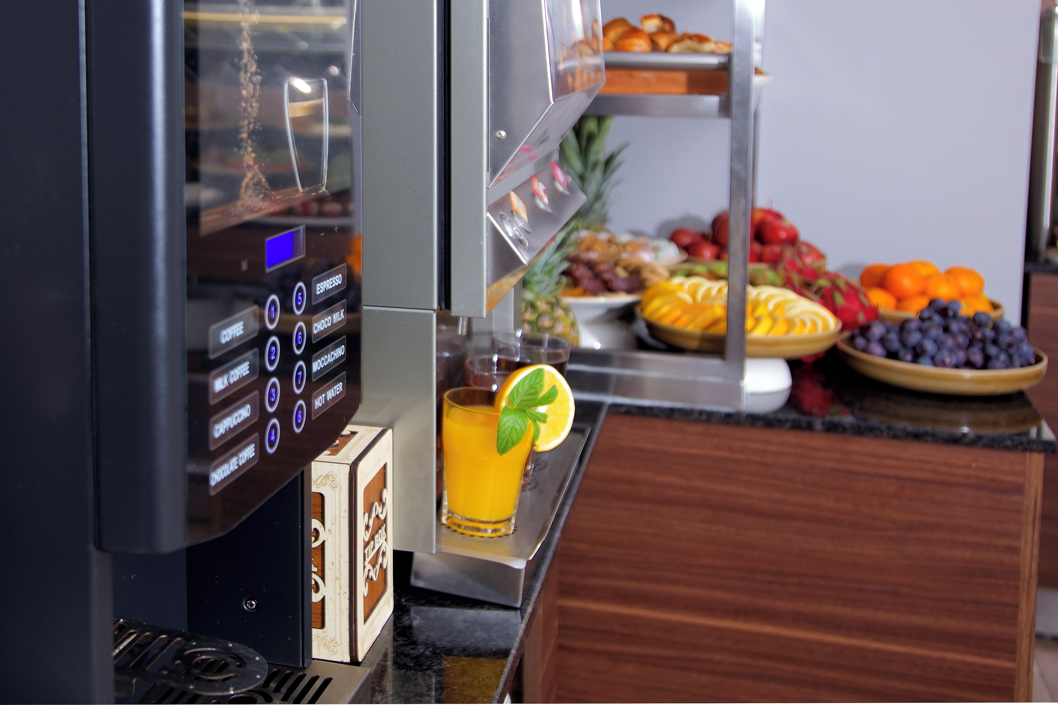 A coffee machine with various drink options, a glass of orange juice with a mint garnish, and a buffet with fruits and pastries.
