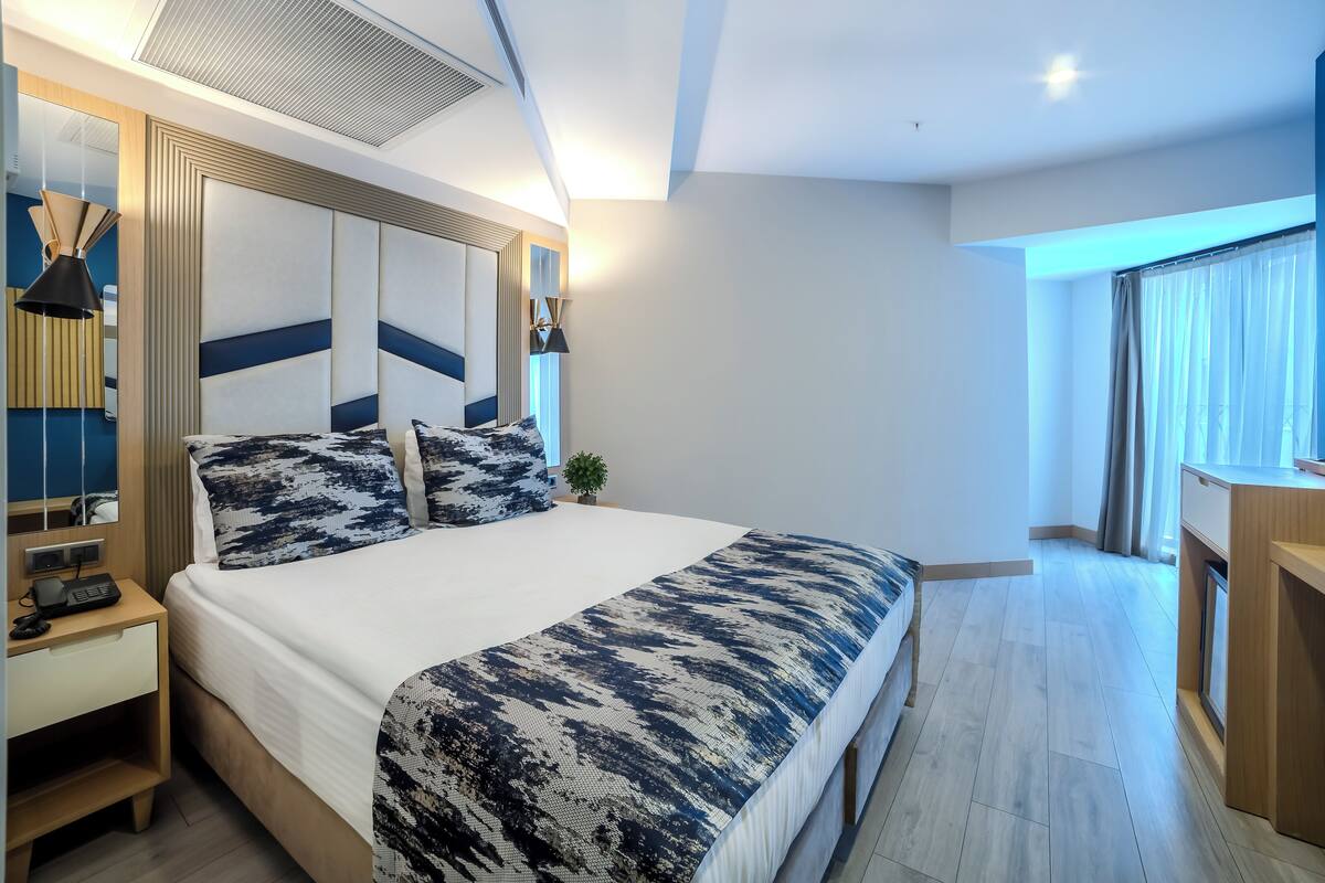 A modern hotel room with a large bed, bedside tables, a desk, and a view of the outside.