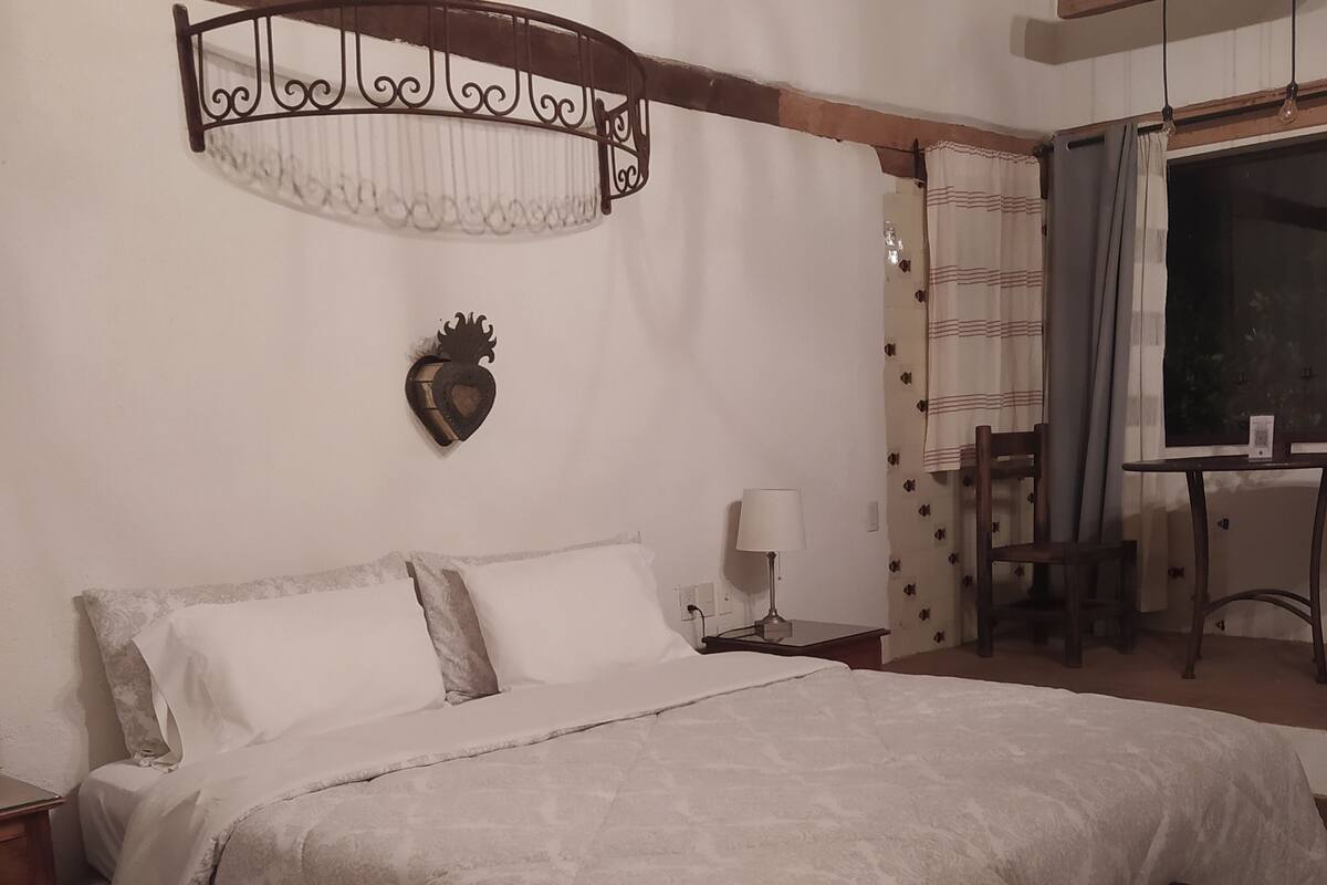 A bedroom with a large bed, a bedside table with a lamp, a wooden bench, a heart-shaped decoration, and a view of the outdoors through a window.