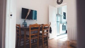 Un comedor con una mesa y sillas de madera, un televisor de pantalla plana montado en la pared, y una cocina visible a través de una puerta abierta.
