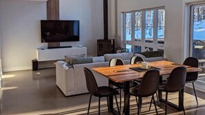 A modern living room with a large TV, a dining table with chairs, and a sofa. - Au Chalet Calme (Lac-Superieur)