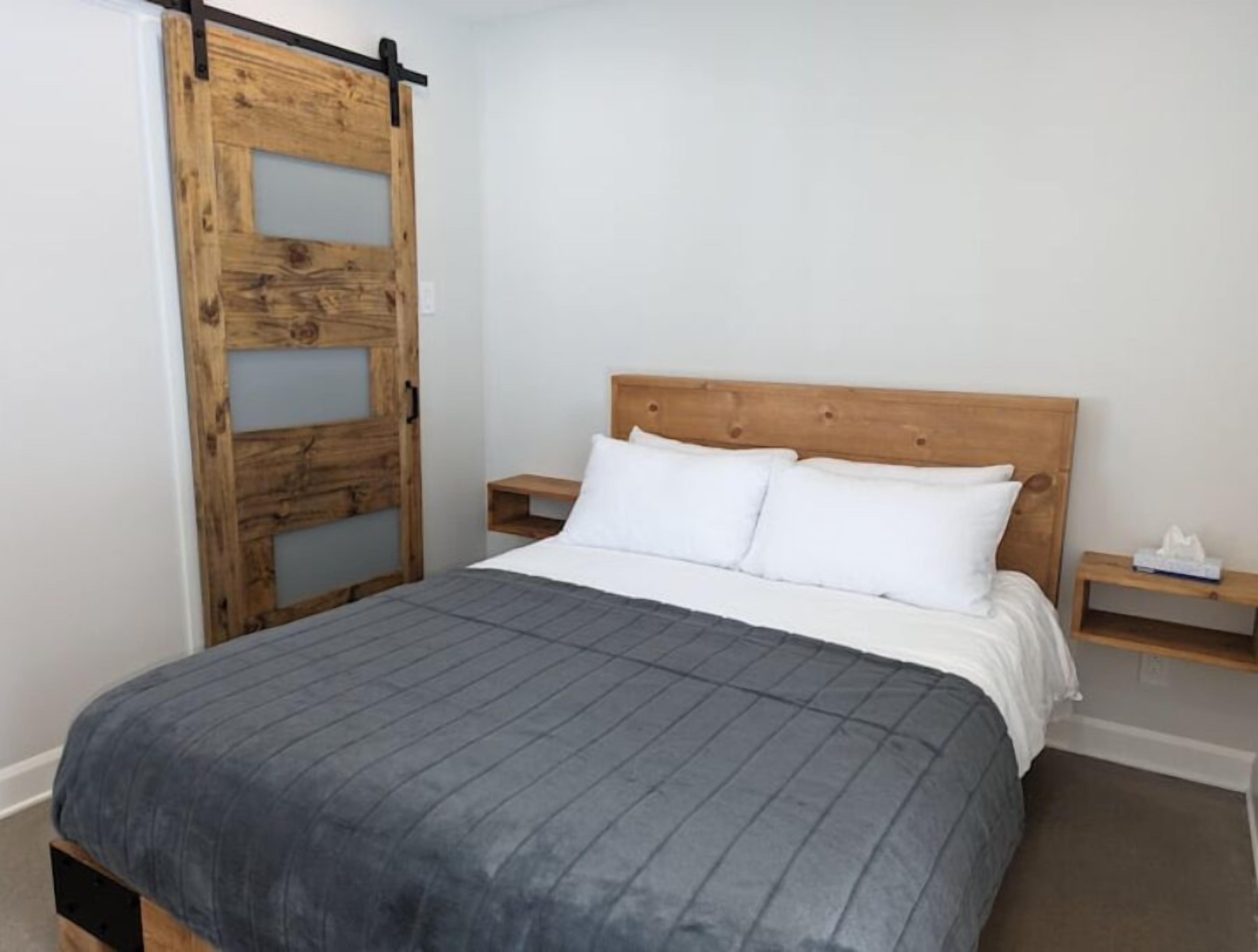 Une chambre à coucher avec un lit en bois, une couvre-lit grise, des oreillers blancs et une table de chevet en bois sur laquelle se trouve une boîte à mouchoirs.