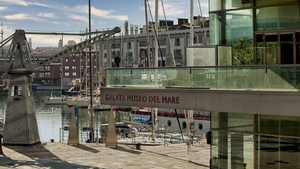 Una marina con imbarcazioni ormeggiate, un ponte sullo sfondo e un edificio con il cartello "GALATA MUSEO DEL MARE".