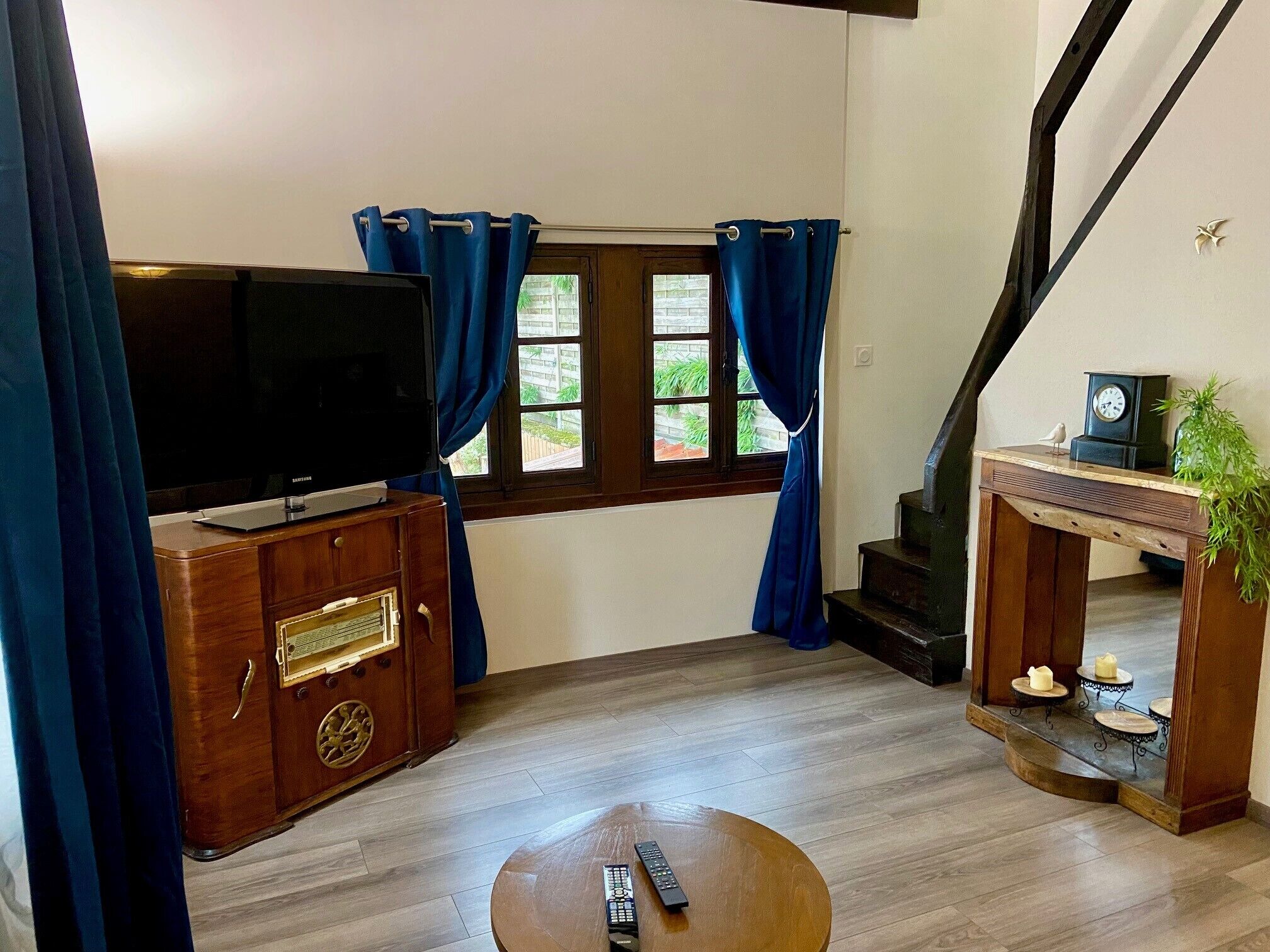 A living room with a TV on a wooden cabinet, a small table with a remote, and a staircase with a clock and plant.