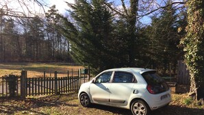 Une voiture blanche est garée dans une zone boisée, avec une clôture en bois et des arbres en arrière-plan.