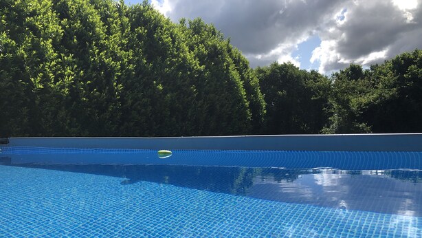 Une piscine d’eau bleue et limpide, avec un objet vert flottant à sa surface, entourée d’un feuillage verdoyant et luxuriant, sous un ciel partiellement nuageux.