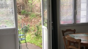 Une pièce dotée d’une porte-fenêtre coulissante donnant sur un jardin. À l’intérieur, on trouve une table en bois sur laquelle sont posés des papiers, ainsi qu’une chaise en métal verte.
