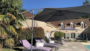 A poolside area with lounge chairs and umbrellas, a stone building in the background, and a palm tree. - Domaine des Rives D'ormoy : Athamante (Ormoy-la-Rivière)