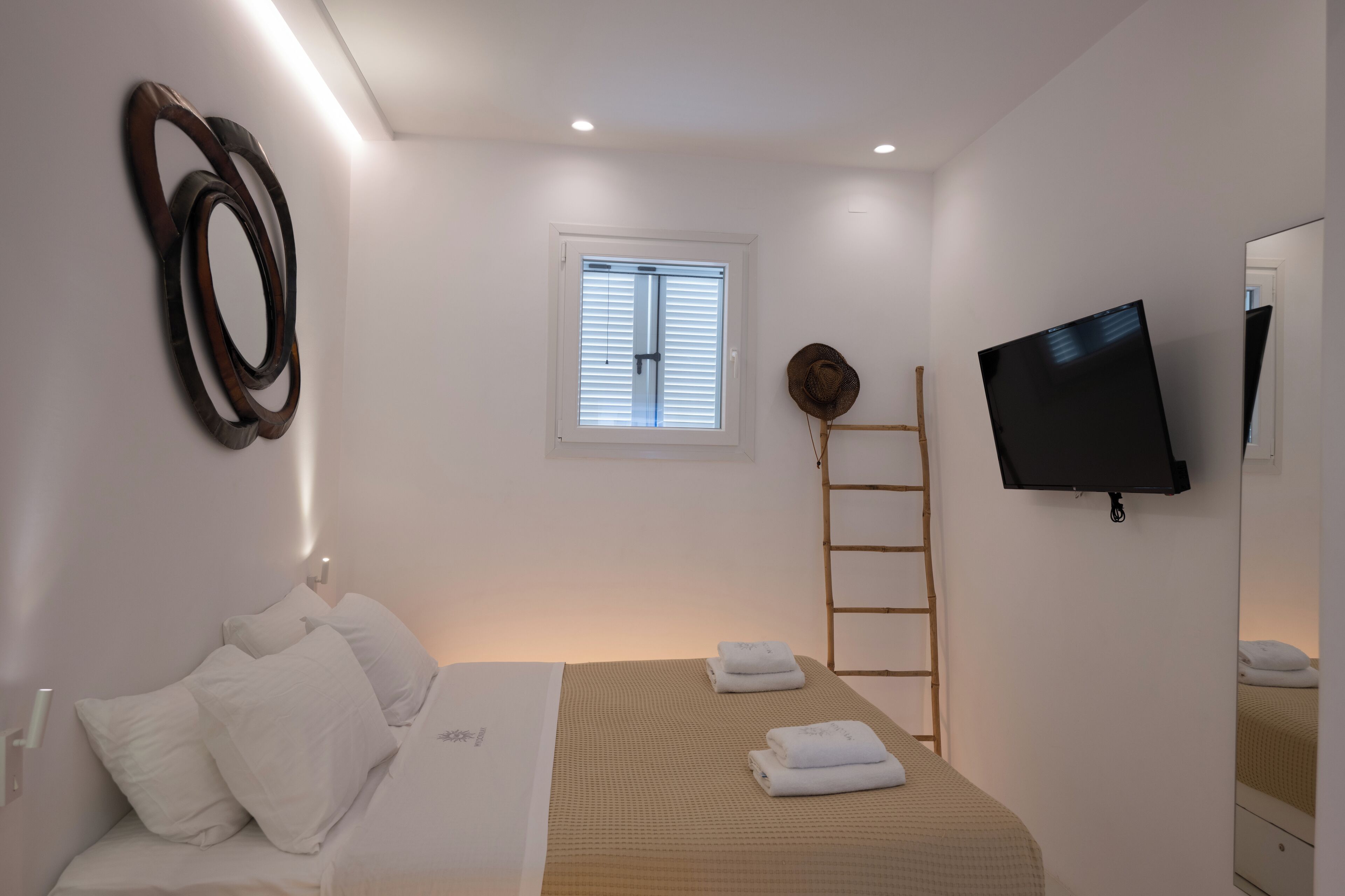 A bedroom with a bed, a TV, a window with blinds, a ladder, and a wall-mounted mirror.
