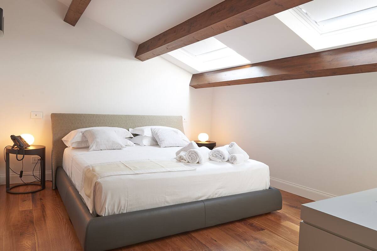 A neatly made bed with white linens and pillows, a bedside table with a lamp, and a wooden floor.