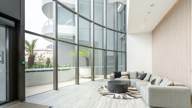 Un hall d’entrée moderne avec un canapé gris, une table basse ronde et un grand mur de verre donnant sur un bâtiment et la verdure environnante.