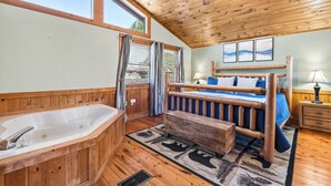 Interior de una cabaña de madera con una bañera grande, una cama con cabecera de madera, un ventilador de techo y una ventana con cortinas.