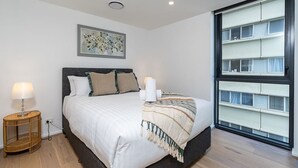 Un dormitorio con una cama grande, una mesita de noche con lámpara, una obra de arte floral enmarcada en la pared y un ventanal con persianas.
