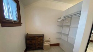 Een kleine, schone kamer met een houten kast, een witte prullenbak en een witte inbouwkast met planken.