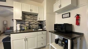 Een moderne keuken met witte kasten, een zwart aanrecht en een magnetron. Aan de muur hangt een brandblusser en er is een klein plankje met een koffiezetapparaat.