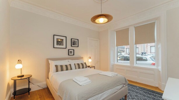 Un dormitorio ordenado con cama, lámparas de noche, ventana con persianas e imágenes enmarcadas en la pared.