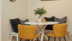 Un comedor con una mesa blanca, dos sillas amarillas, cojines estampados en blanco y negro, un reloj, una obra de arte enmarcada y una lámpara de pie.