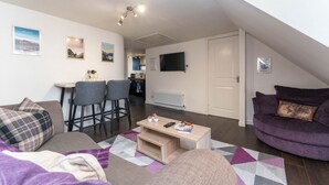 A modern loft apartment with a dining area, a living room with a purple sofa, and a kitchen in the background. - Perth Penthouse at the Concert Hall (Perth and Kinross)
