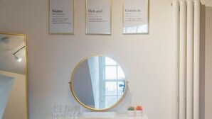 A bedroom with a white vanity, a round mirror, a pink bench, and a bed with a plaid blanket. - Perth Penthouse at the Concert Hall (Perth and Kinross)
