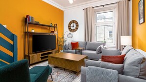 Una sala de estar con sofá, mesa de centro y televisor, una estantería y un reloj en la pared.