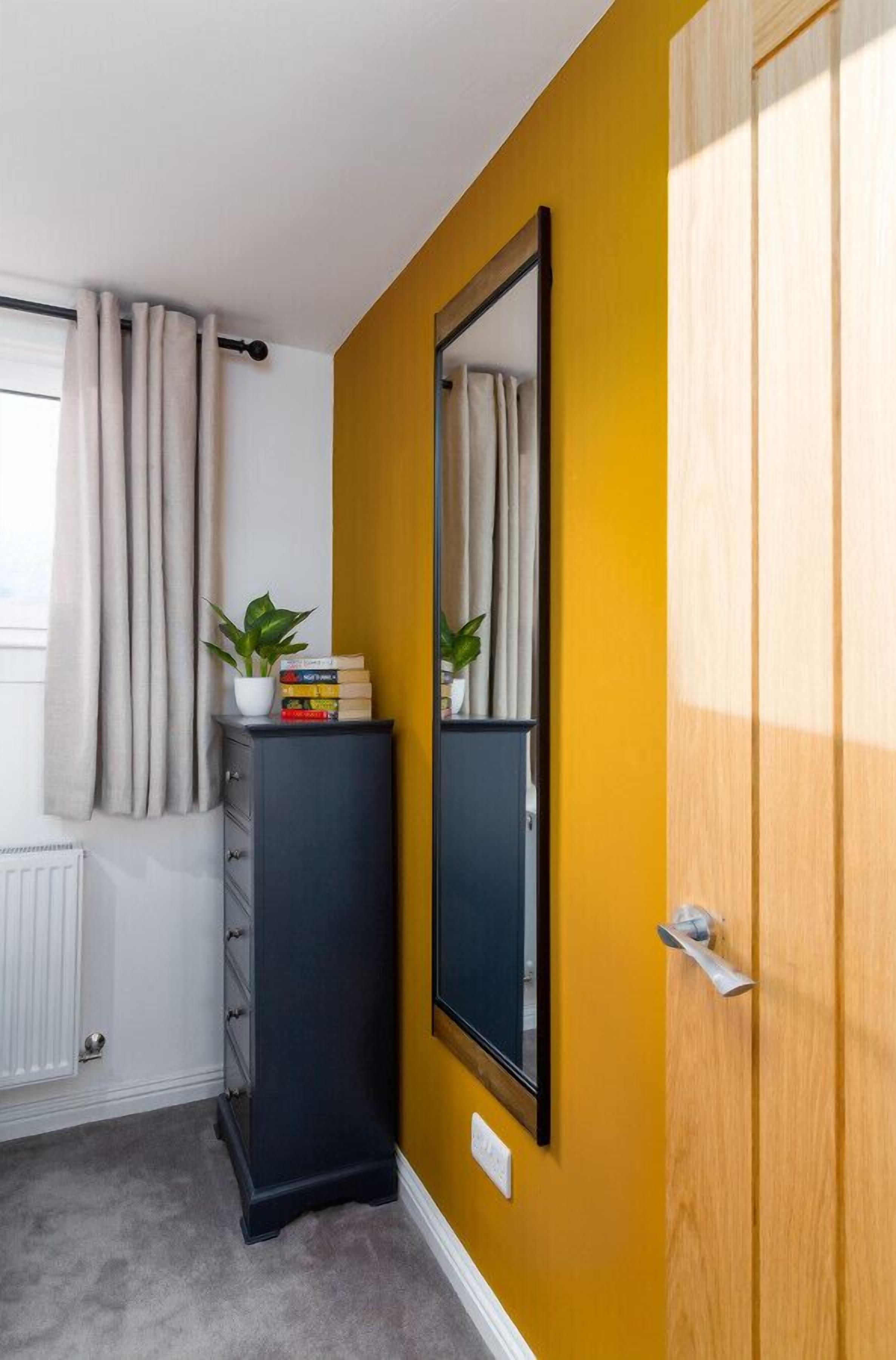 Une pièce avec un mur jaune, un grand miroir, une porte en bois, un radiateur, un meuble noir et une fenêtre avec des rideaux.