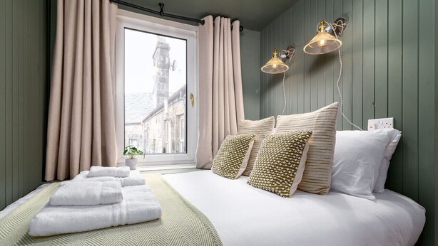 Una cama bien hecha con sábanas blancas y almohadas estampadas, una ventana con cortinas y dos lámparas fijadas a la pared.