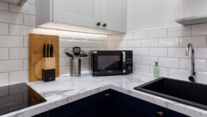 Una cocina moderna con azulejos blancos tipo metro, un microondas negro, una tabla de cortar de madera y un lavamanos de acero inoxidable.
