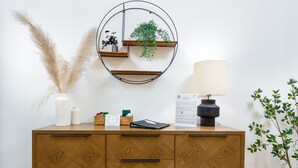 Un armario de madera con cajones, un estante redondo montado en la pared con plantas, una lámpara y una pequeña planta en maceta a su lado.