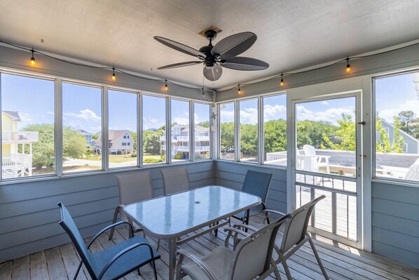 A covered patio with a glass table and chairs, large windows, and a ceiling fan. - Sun Realty oceanside classic in the heart of Duck, NC (Duck)