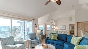 A spacious living area with a large window, a ceiling fan, and a blue sofa. - Oceanside vacation rental steps away from the beach access in Corolla, NC! (Corolla)