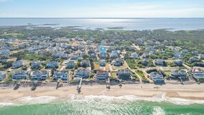 An aerial view of a coastal residential area with houses along the beachfront, a pier extending into the water, and a clear view of the ocean and distant land. - Oceanside vacation rental steps away from the beach access in Corolla, NC! (Corolla)