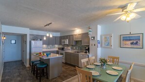 A modern kitchen with a central island, bar stools, a dining table set for four, and a ceiling fan. - Beautiful sunrise views from this oceanfront condo in Kill Devil Hills NC! (Kill Devil Hills)