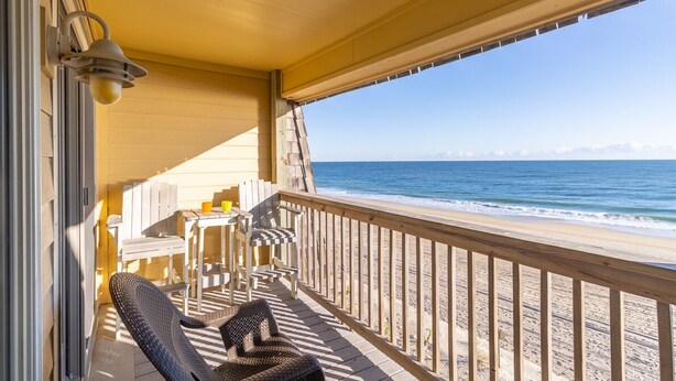 A covered outdoor area with a wooden deck, chairs, and a table. - Beautiful sunrise views from this oceanfront condo in Kill Devil Hills NC! (Kill Devil Hills)