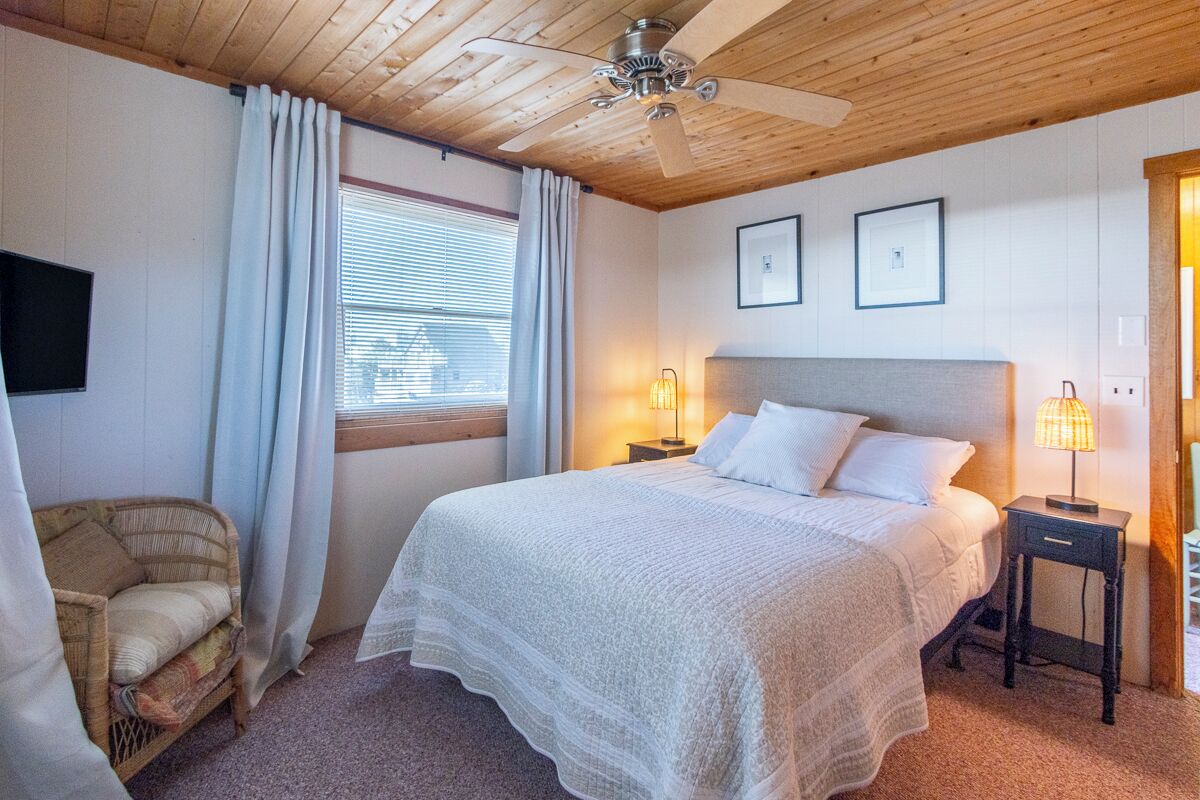 Une chambre avec un grand lit, un ventilateur de plafond, une fenêtre avec des rideaux, une table de chevet avec une lampe et une chaise.