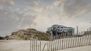A beachfront house with multiple balconies, a sandy beach in the foreground, and a cloudy sky.