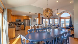 A spacious kitchen with wooden cabinets, a dining area with blue chairs, and a large window overlooking the ocean.