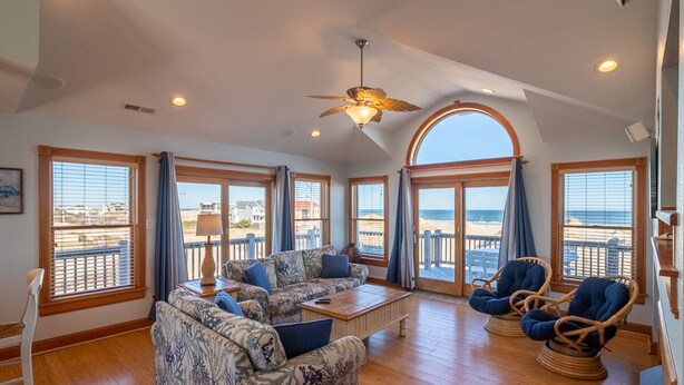 A spacious living room with a large sofa, two armchairs, and a coffee table. The room has large windows with ocean views and a ceiling fan.