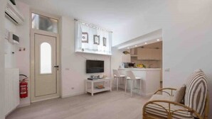 Een compacte leefruimte met een kitchenette, een televisie, een eetgedeelte en een zithoek met een rieten stoel en een bank.