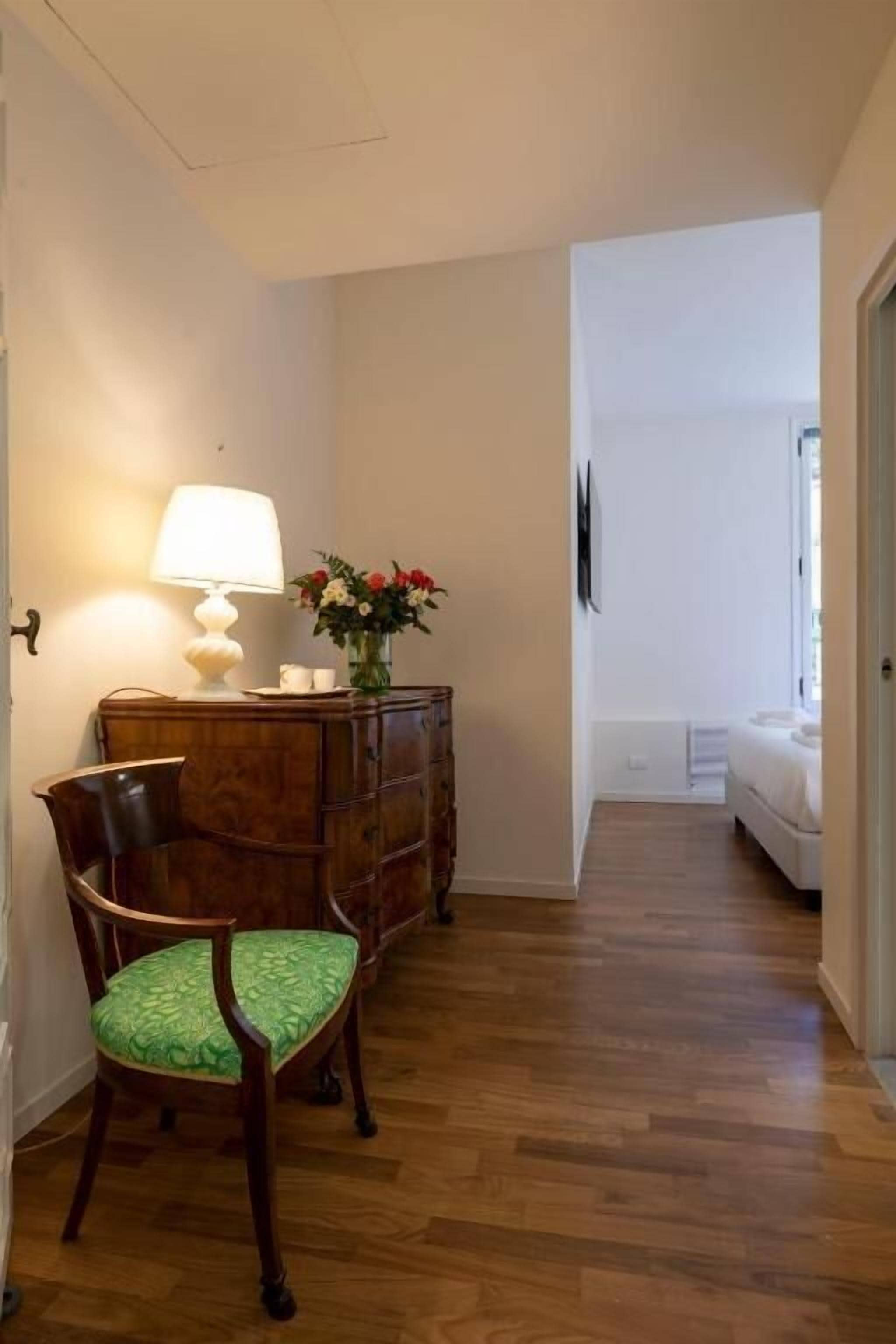 A hallway with a wooden floor, a wooden chair with a green cushion, a wooden dresser with a lamp, and a vase with flowers.