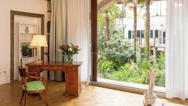A room with a wooden desk, a chair, a lamp, a vase with flowers, and a view of a garden through a large window. - Mocenigo Grand Canal Luxury Suites (Venice)